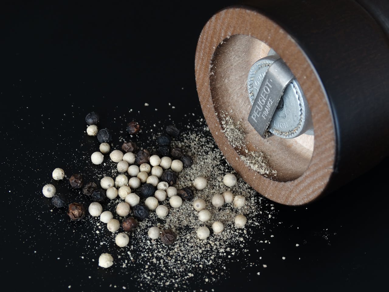 Close-up of a pepper grinder and assorted peppercorns on black surface.